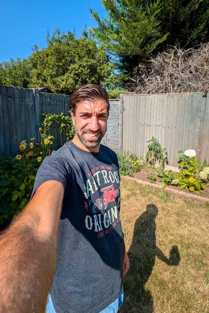 Man taking a selfie in a garden with plants and a wooden fence.